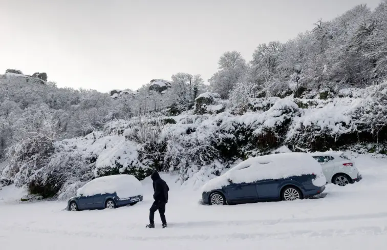 Pessoa caminha na neve, onde estão carros estacionados cobertos de neve.